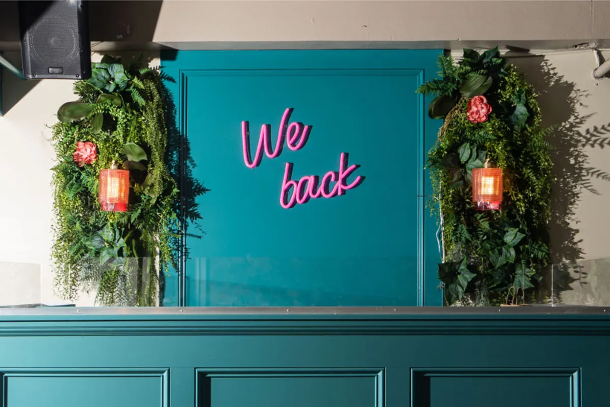 A playful interior scene featuring a neon sign reading 'We back', lush greenery, and decorative lighting. The space buzzes with social gatherings and captures the spirit of vibrant nightlife.