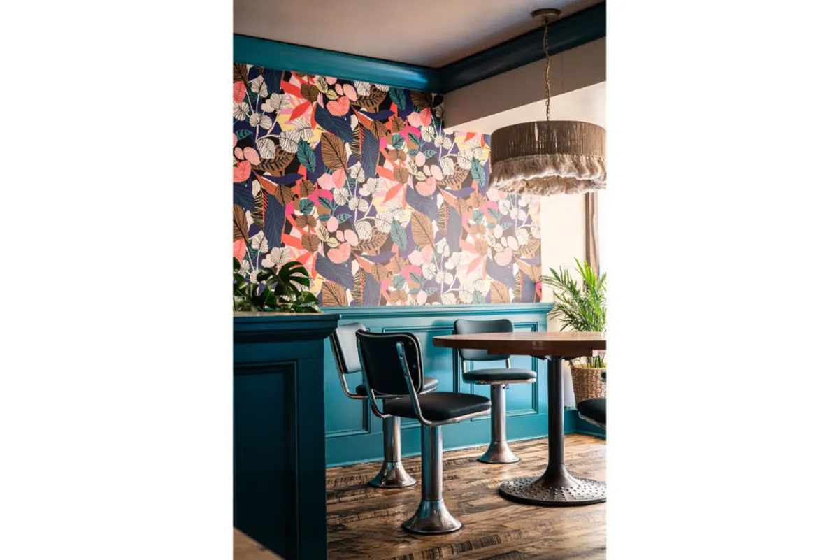 A vibrant living space with creative floral wallpaper, contemporary pendant lighting, a round wooden table, and stylish black chairs. The atmosphere is cheerful and modern, ideal for working or relaxing, echoing the room's dual purpose.