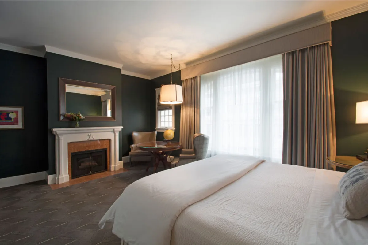 This image showcases a plush bedroom featuring a king-size bed, a glowing gas fireplace, expansive windows bringing in natural light, a mirror, and a cozy seating area. The decor feels elegant and welcoming, with subtle cultural and artistic touches visible in the arrangement.