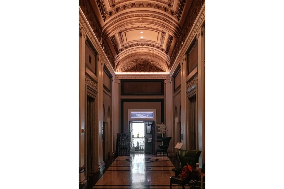 A sophisticated, welcoming hallway with ornate ceiling moldings, a stylish green armchair, and a doorway at the far end. The architectural details and artful layout invite guests to wander and discover, suggesting both comfort and the anticipation of new experiences.