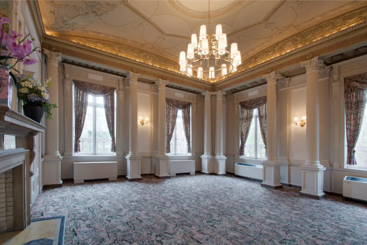 A spacious and sophisticated interior featuring a magnificent chandelier, ornate ceiling, large windows with flowing curtains, and a grand fireplace. The scene feels open and inviting, with ample space that could host art exhibitions or community gatherings. Decorative elements and artwork contribute to an atmosphere of creative inspiration and refined design.