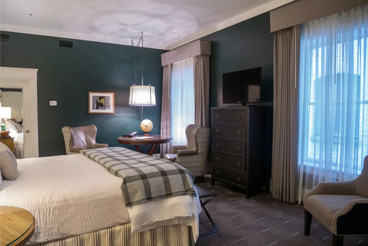 The image showcases a bright, elegantly furnished Premier Corner King room. A plush bed with a gray checkered throw is the centerpiece, complemented by two stylish armchairs and a round wooden table with a warm lamp. Large windows fill the room with natural light, creating an inviting, comfortable retreat perfect for relaxing, reading, or enjoying a quiet glass of wine.