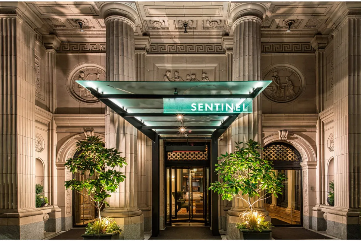 A sophisticated entrance to the Sentinel hotel, featuring an elegant awning with the 'SENTINEL' sign, impressive architectural columns, and lush potted plants flanking the doorway. Guests are seen gathering or socializing at the entry, creating an inviting atmosphere.