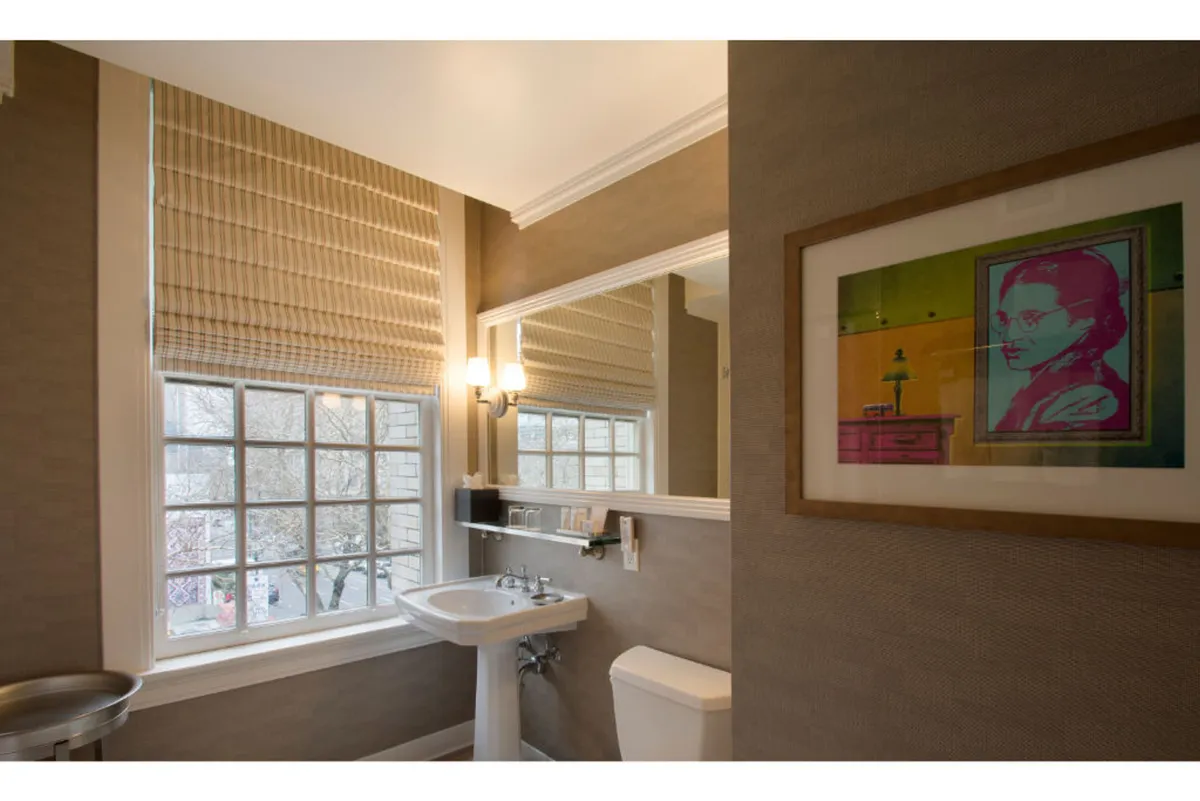 A serene space with a window shaded by blinds, curated artwork on the wall, a sleek sink, and mirror. The area is bathed in gentle natural light, evoking inspiration and calm—a nod to the room’s artistic and contemplative elements.
