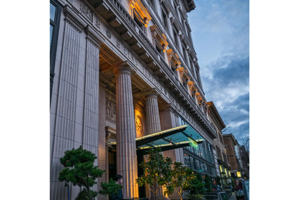 A street-level view of Sentinel’s illuminated facade at dusk, showcasing ornate columns, a glass entrance canopy, and greenery that frames the entrance. The scene captures the lively ambiance of the surrounding neighborhood, ideal for evening strolls or dining out.