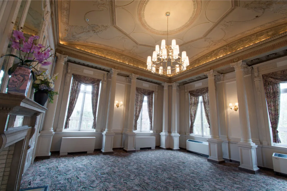 An elegant interior with a brilliant chandelier, intricate ceiling details, tall windows that flood the room with natural light, and tasteful floral arrangements. The setting feels both luxurious and artistic, ideal for intimate gatherings and art showcases.